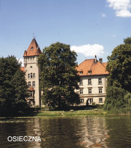 Pałac w Osiecznej otoczony stawem i drzewami widziany w dzień. Budynek w jasnym kolorze wielokondygnacyjny z czerwonym dachem.